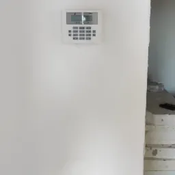 Biały panel sterowania alarmem z klawiaturą i wyświetlaczem LCD, zamontowany na świeżo pomalowanej białej ścianie w pomieszczeniu w trakcie remontu, widoczne elementy wykończeniowe i podłoga w tle.