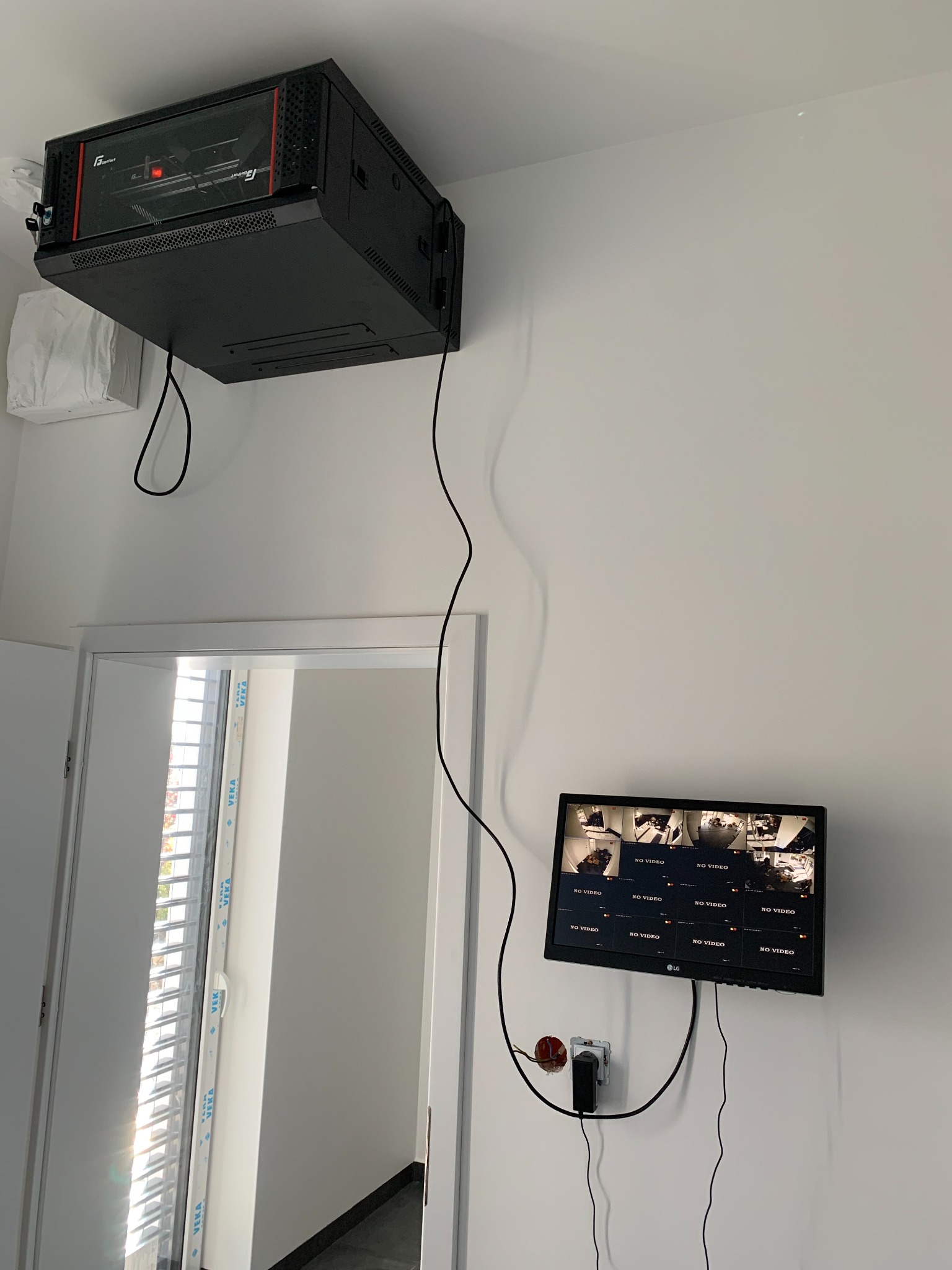 Szafa serwerowa rack zamontowana pod sufitem w pomieszczeniu z białymi ścianami, obok drzwi balkonowych i monitora z podglądem z kamer, widoczne kable i gniazdko elektryczne z zasilaczem.