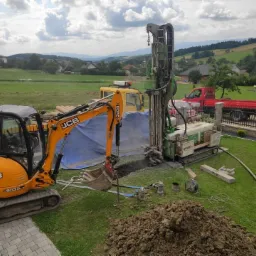 Żółta koparka JCB B030 w trakcie prac ziemnych obok wiertnicy na gąsienicach; w tle widoczny krajobraz wiejski i inne pojazdy.