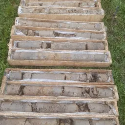 Rdzenie glebowe ułożone w drewnianych skrzynkach z numeracją, zabezpieczone folią, leżące na trawie.