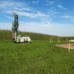 Wiertnica na gąsienicach, ustawiona na trawiastym terenie z wysoką kukurydzą w tle, pod błękitnym niebem z chmurami. Przygotowane drewniane konstrukcje w pobliżu sugerują początek prac budowlanych.