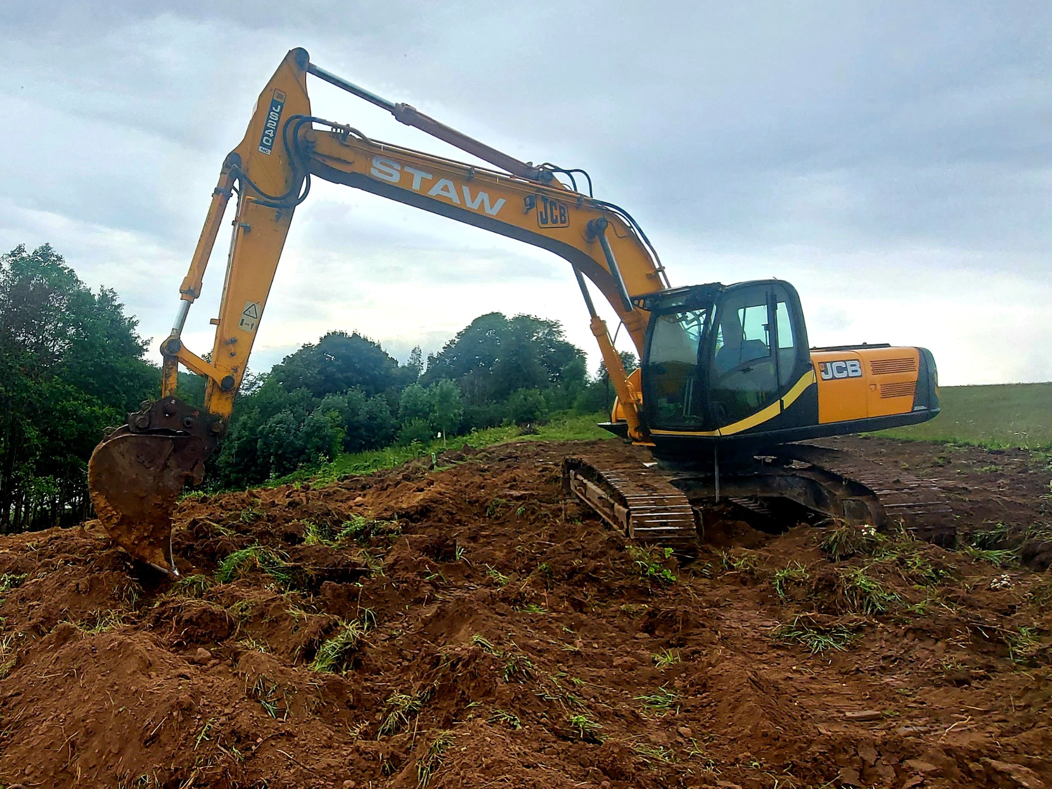 Żółto-czarna koparka JCB z łyżką na nierównym terenie z rudą ziemią, w tle drzewa i pochmurne niebo, widoczny napis 'STAW' na ramieniu koparki.