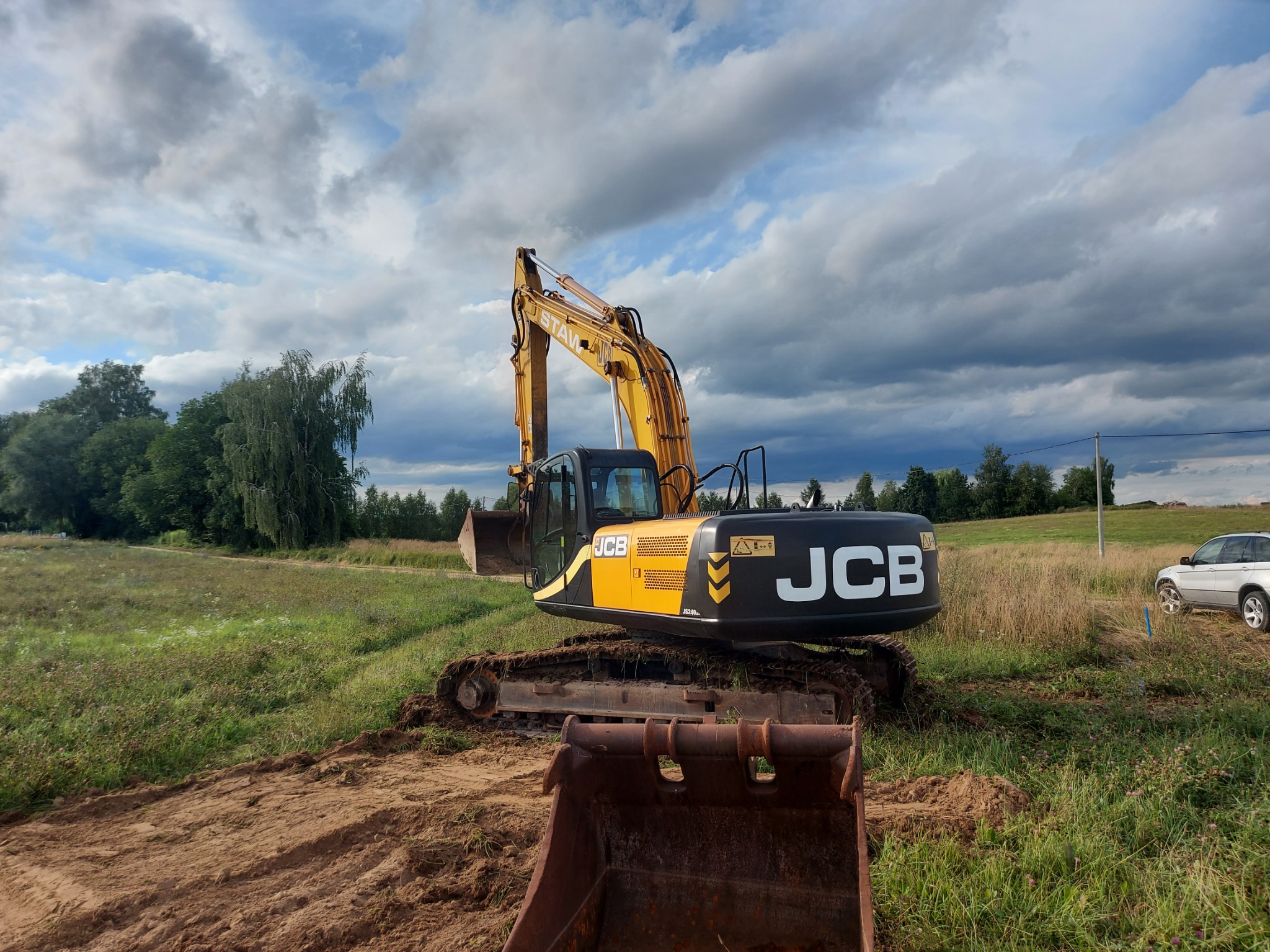 Żółto-czarna koparka JCB na polu z wykopaną ziemią i widocznym drugim wiadrem na pierwszym planie, w tle drzewa, pochmurne niebo i samochód.