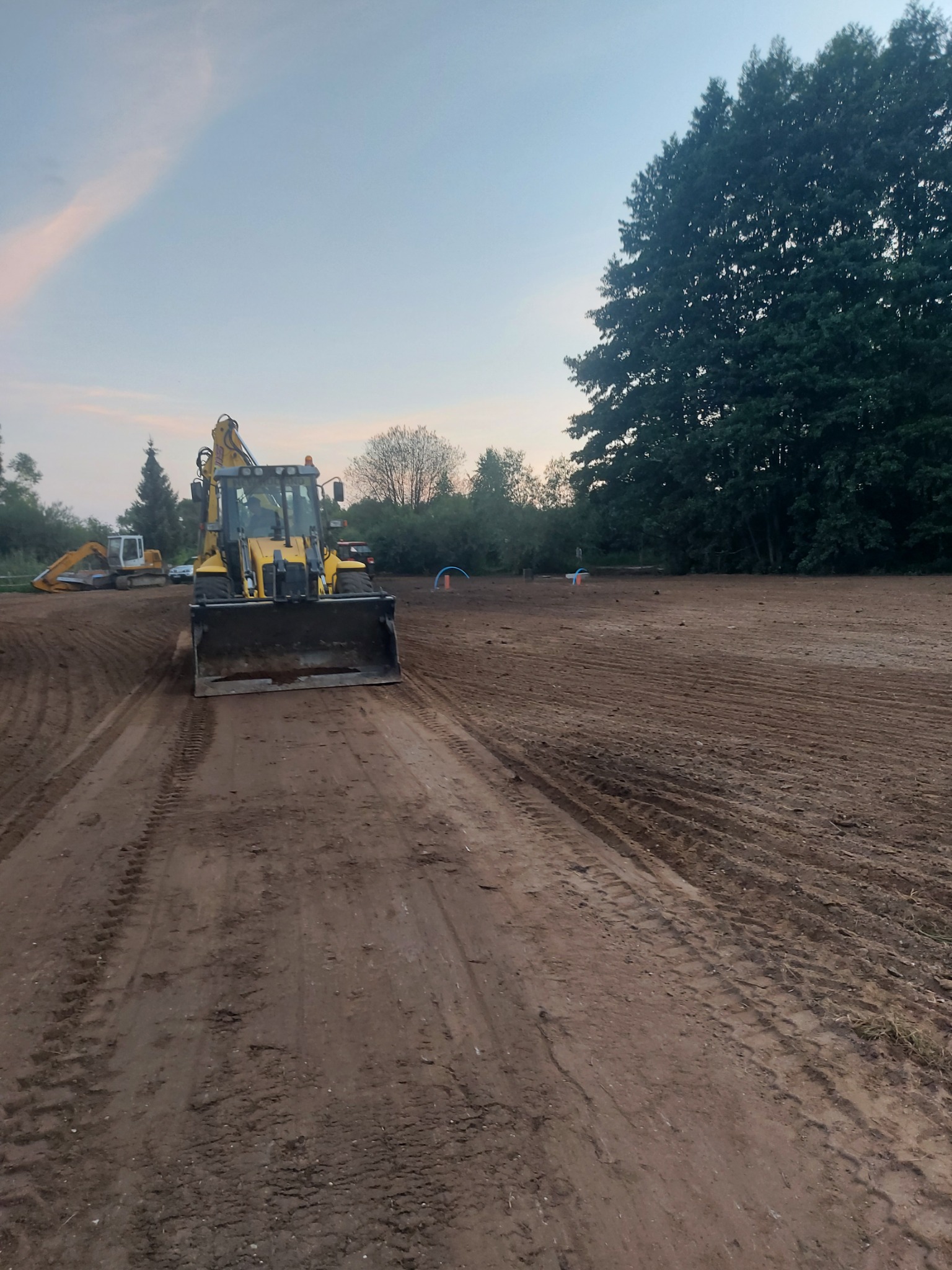 Żółta koparko-ładowarka na przygotowanym terenie, widoczne ślady opon i wyrównana ziemia, w tle drzewa i zachmurzone niebo.