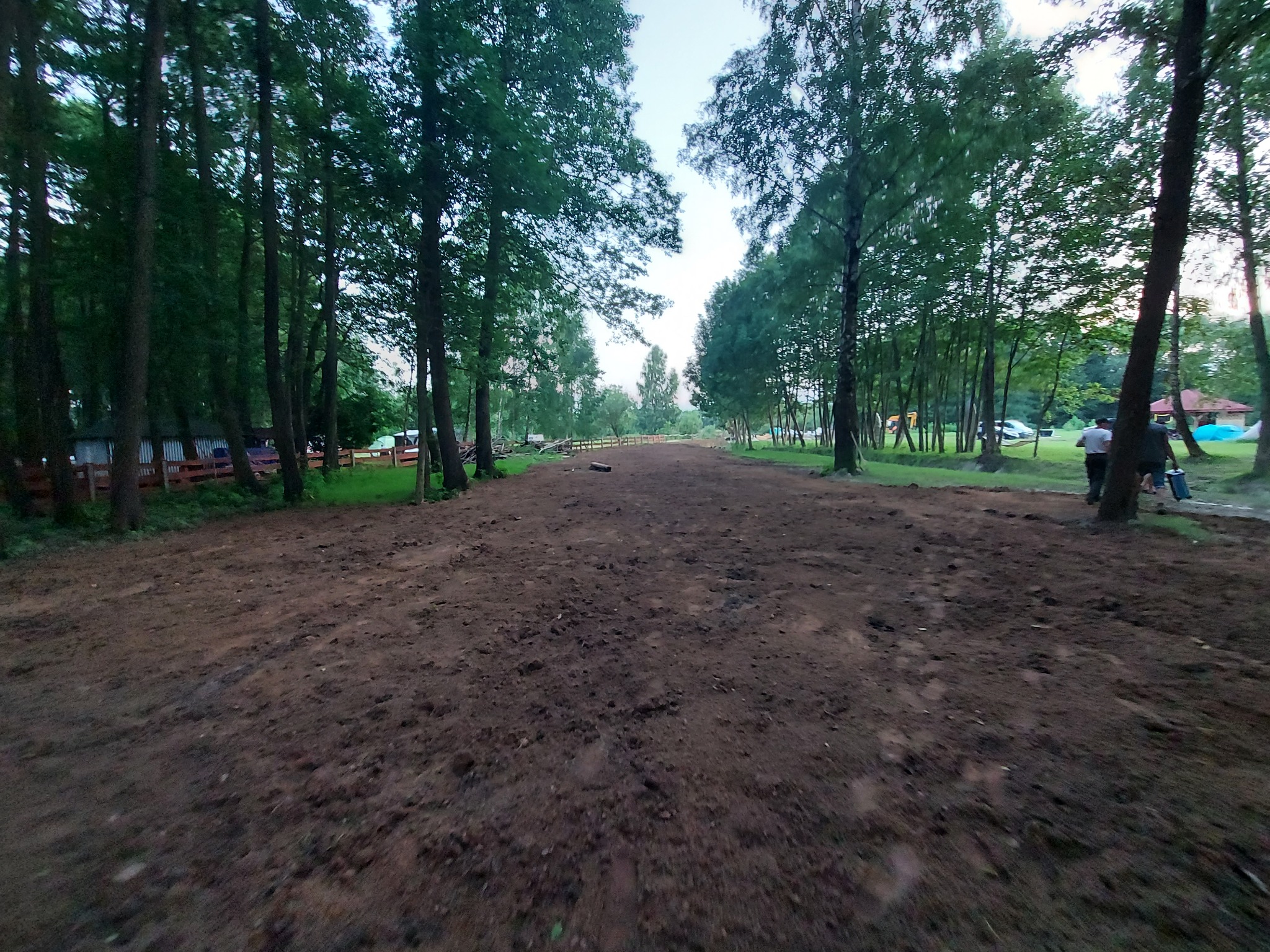 Udrożniona droga gruntowa wśród drzew, ślady opon widoczne na powierzchni, w tle zabudowania i sylwetki ludzi.