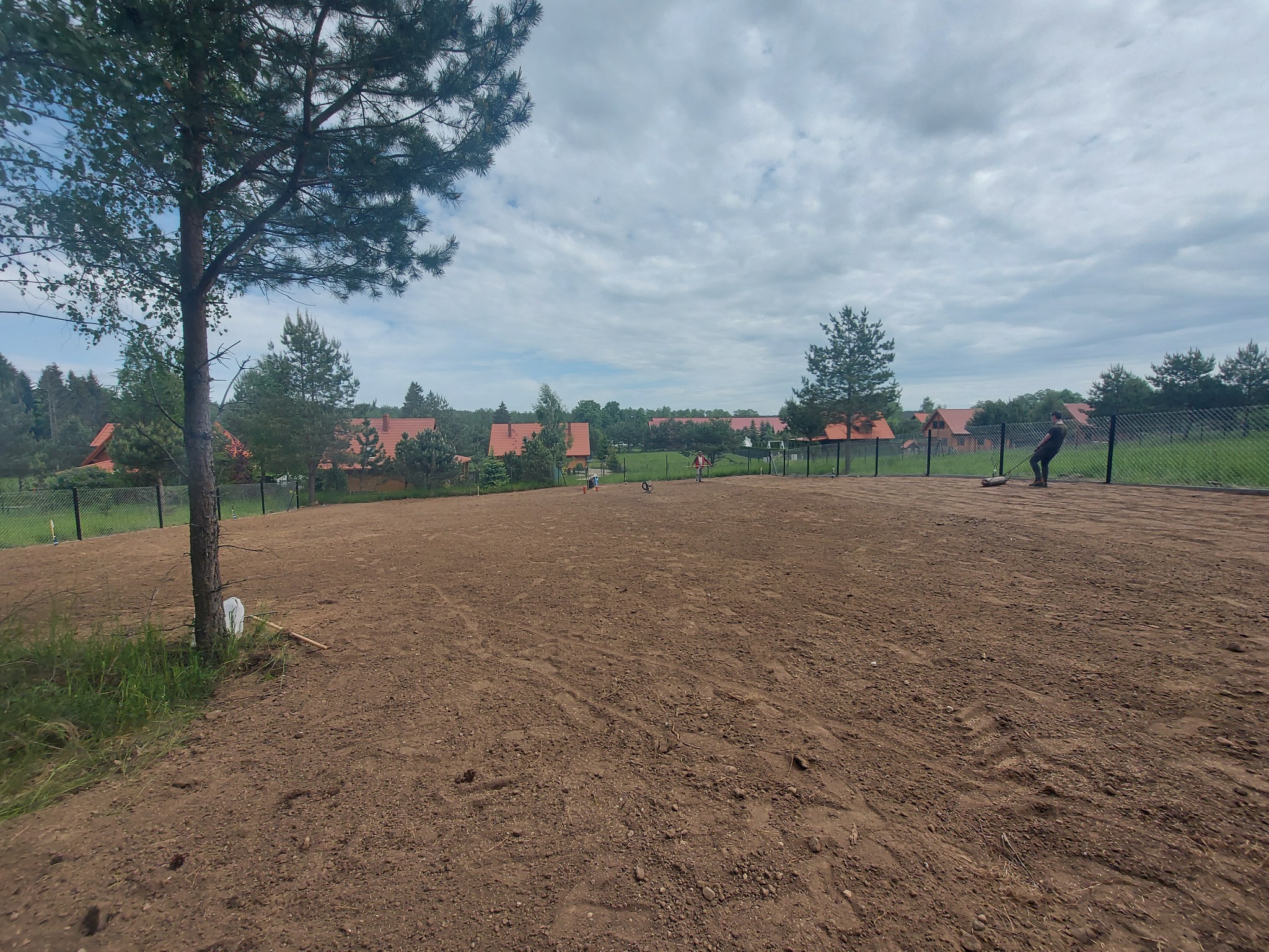 Przygotowany teren pod budowę z widoczną świeżo wyrównaną ziemią, ogrodzeniem z siatki i osobą grabiącą w tle, otoczony drzewami i domami z czerwonymi dachami.