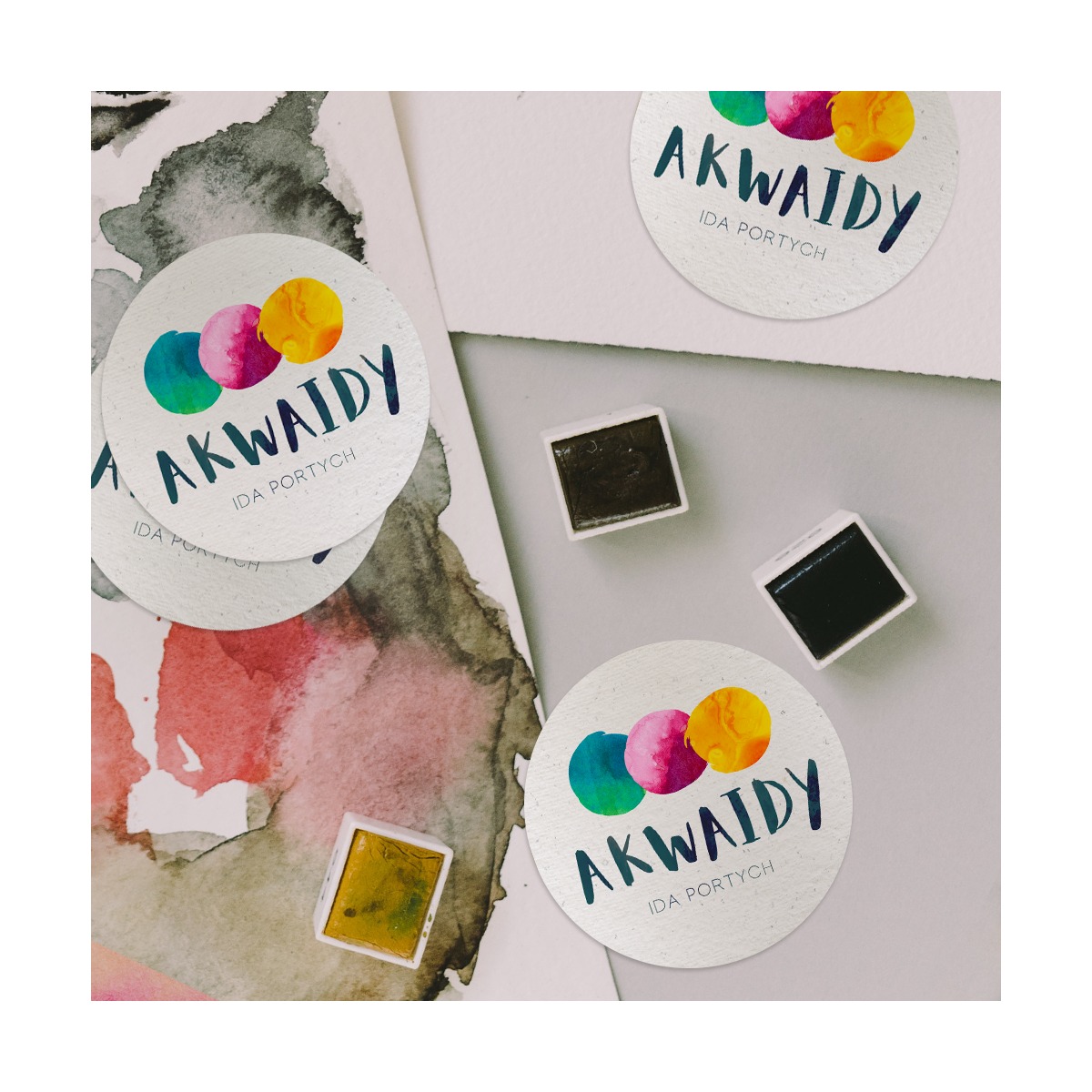 Sticker z logo 'AKWAIDY IDA PORTYCH' z trzema kolorowymi kropkami, obok próbki farb akwarelowych i fragment akwareli na papierze.