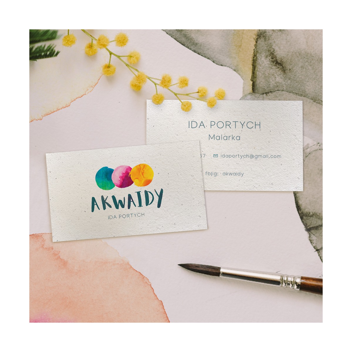 Dwie wizytówki z logo firmy malarskiej 'Akwaidy' na tle akwareli i pędzla, udekorowane gałązką mimozy.