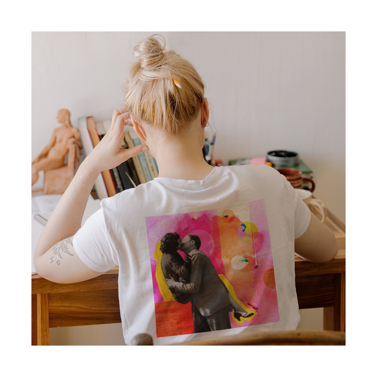Kobieta siedzi przy biurku, tyłem, w białej koszulce z nadrukiem kolażu przedstawiającego całującą się parę na tle abstrakcyjnych kształtów i kolorów.