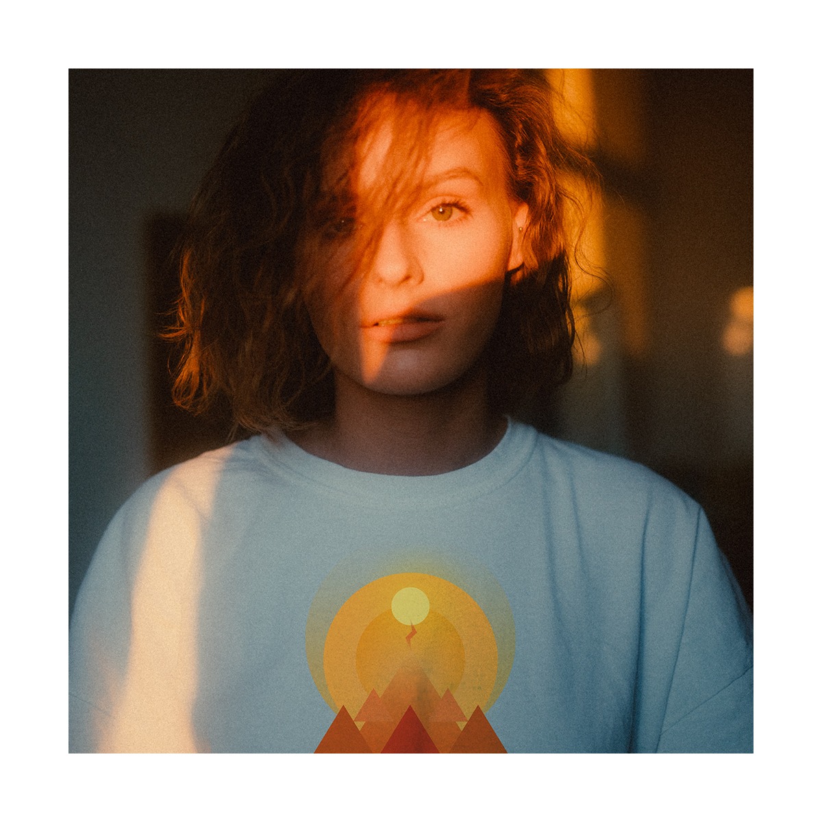 Portret kobiety z włosami w odcieniach rudości, oświetlonej ciepłym, zachodzącym słońcem, ubrana w białą koszulkę z graficznym nadrukiem przedstawiającym góry i słońce.