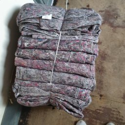 Transport przeprowadzki - Stos szarych koców transportowych z kolorowymi włóknami, spiętych białym sznurkiem, leżących na brudnej drewnianej podłodze obok pary białych butów.