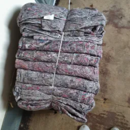 Stos szarych koców transportowych z kolorowymi włóknami, spiętych białym sznurkiem, leżących na brudnej drewnianej podłodze obok pary białych butów.