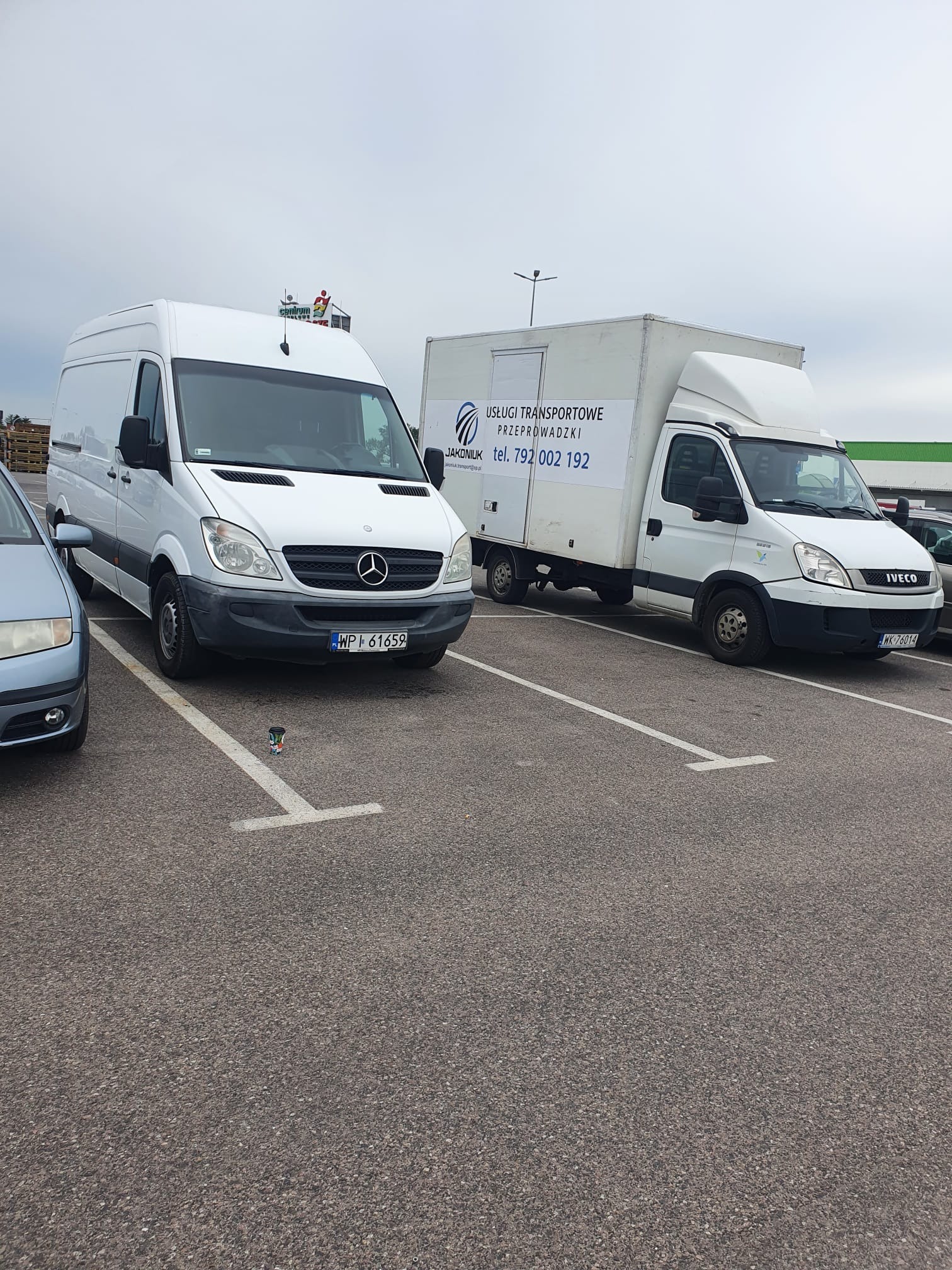 Dwa białe busy transportowe zaparkowane na parkingu, jeden z reklamą usług transportowych i przeprowadzkowych, obok kubek po kawie