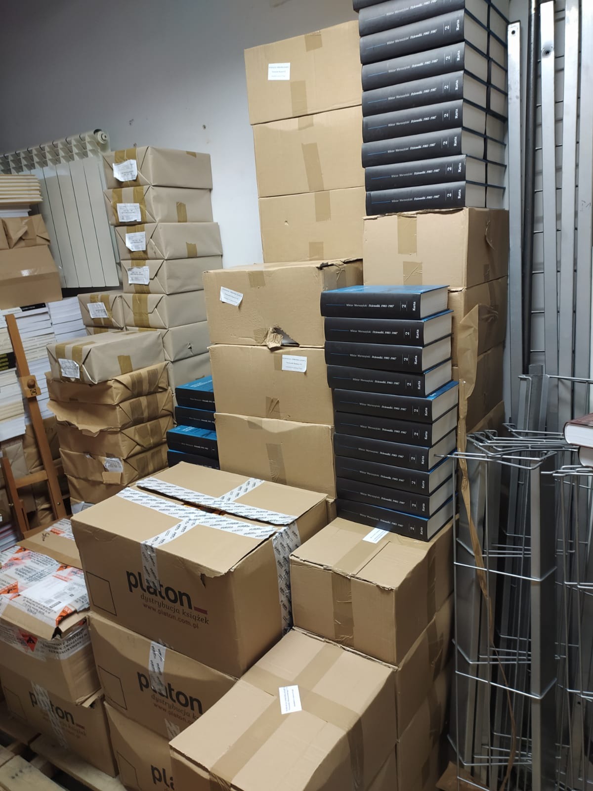 Stosy zapakowanych książek i kartonów, niektóre z logo 'platon dystrybucja książek', przygotowane do transportu w magazynie.