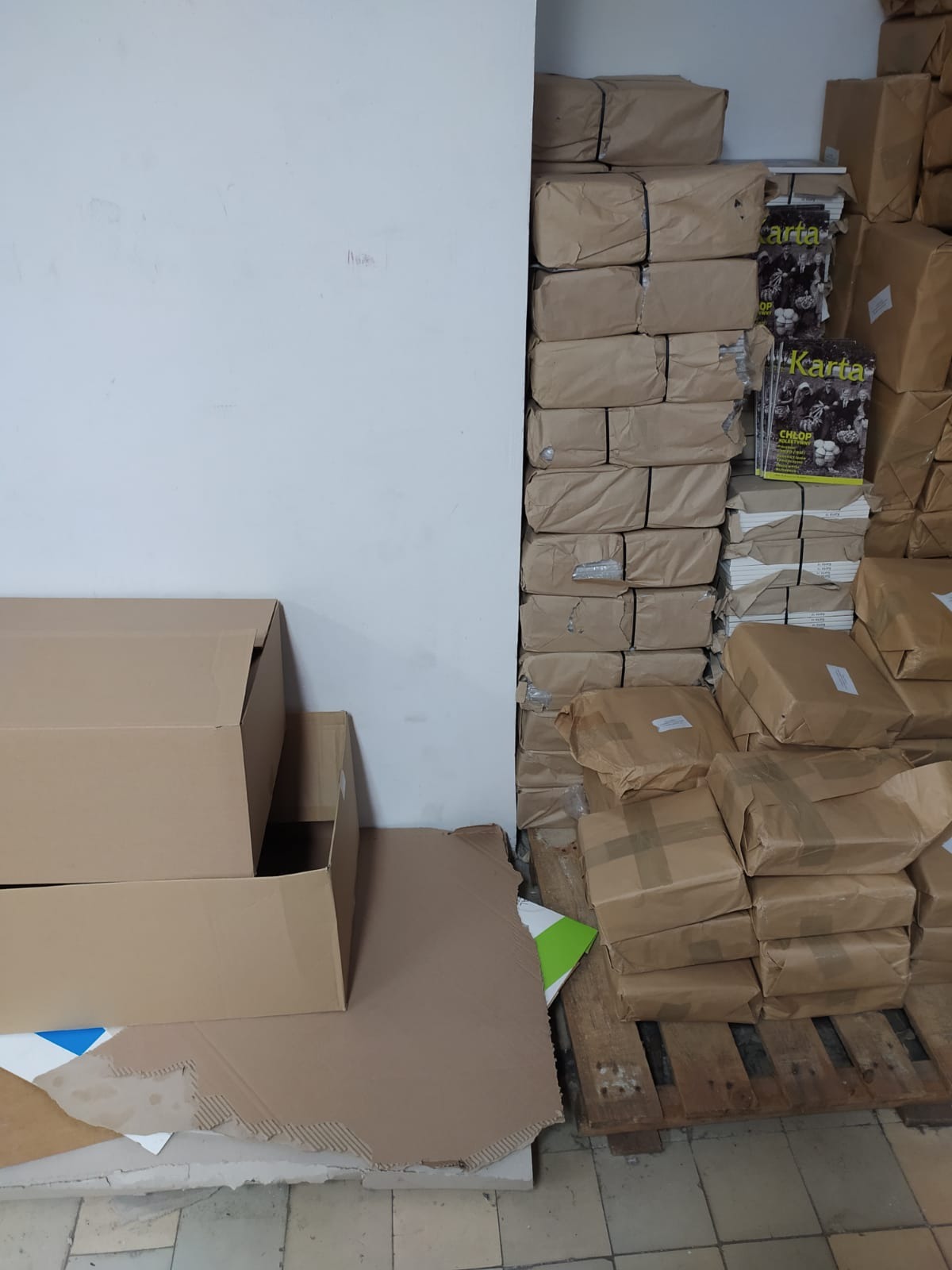 Stos zapakowanych paczek na drewnianej palecie obok białej ściany, częściowo widoczne kartony i magazyn 'Karta'.