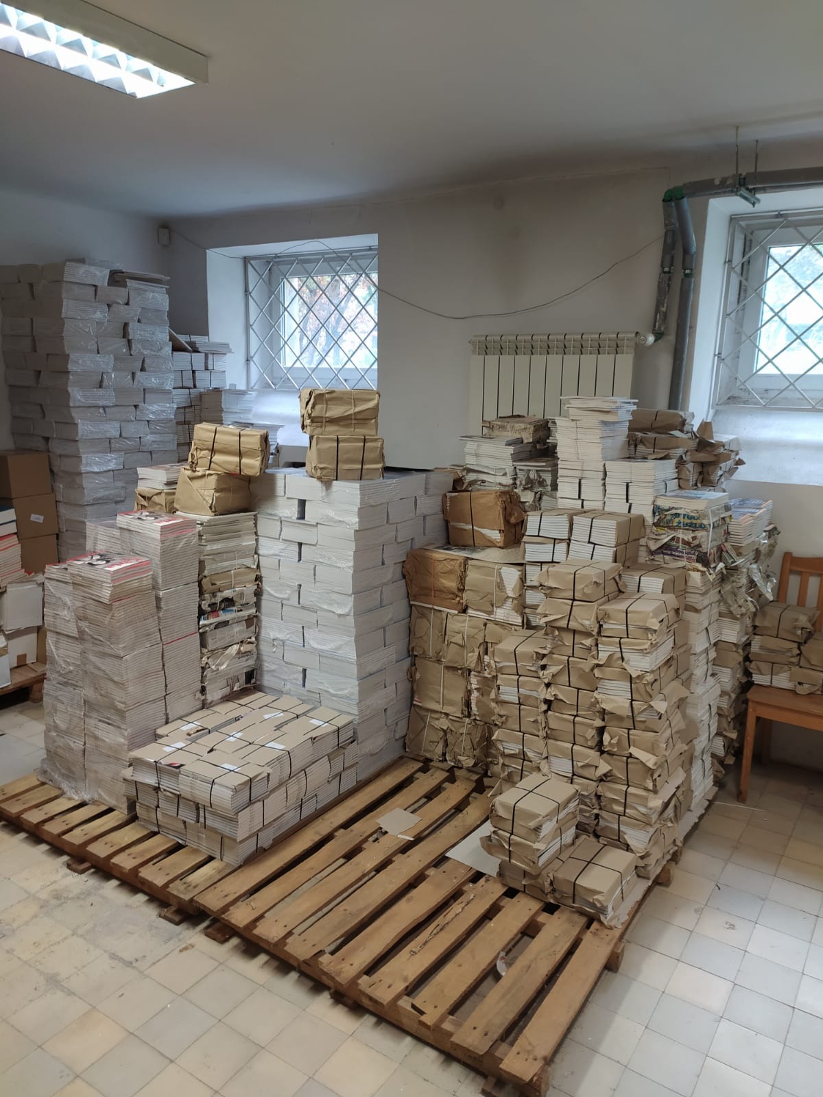 Stosy zapakowanych książek i gazet na paletach w magazynie z oknami w kratkę i grzejnikiem.