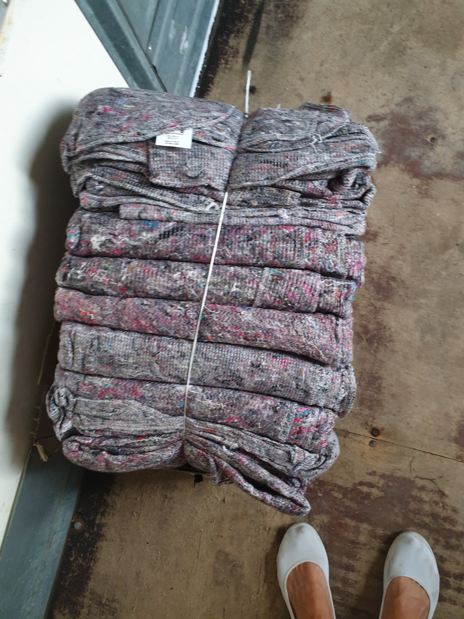 Stos szarych koców transportowych z kolorowymi włóknami, spiętych białym sznurkiem, leżących na brudnej drewnianej podłodze obok pary białych butów.
