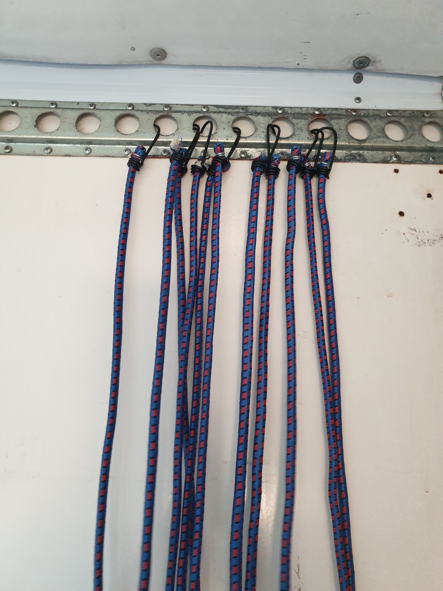 Siedem elastycznych, niebiesko-czerwonych lin z haczykami, zawieszonych na metalowej szynie z otworami, przymocowanej do białej ściany wewnątrz pojazdu.