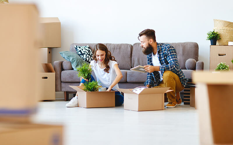 Uśmiechnięta para pakuje rośliny doniczkowe i ramkę ze zdjęciem do kartonowych pudeł podczas przeprowadzki, na podłodze i w tle widoczne inne zapakowane kartony.