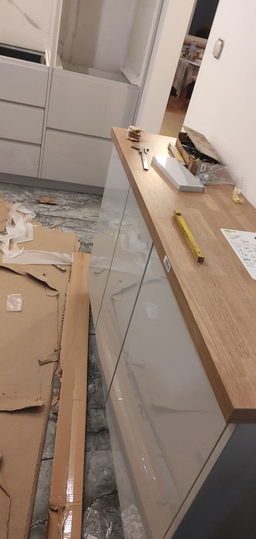 Montaż blatu drewnianego na szafkach kuchennych z białymi frontami imitującymi marmur, widoczne narzędzia i materiały montażowe na blacie, podłoga zabezpieczona kartonem i folią.