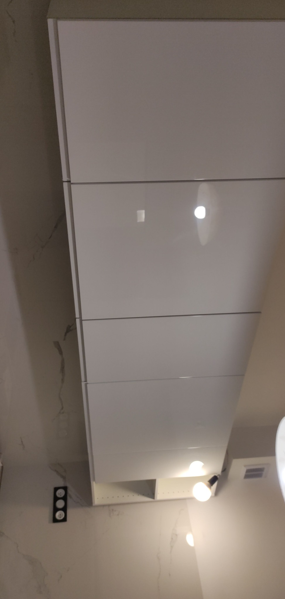 Białe, lakierowane fronty szafek kuchennych pod sufitem, widoczne oświetlenie punktowe i marmurowa ściana z gniazdkami elektrycznymi.