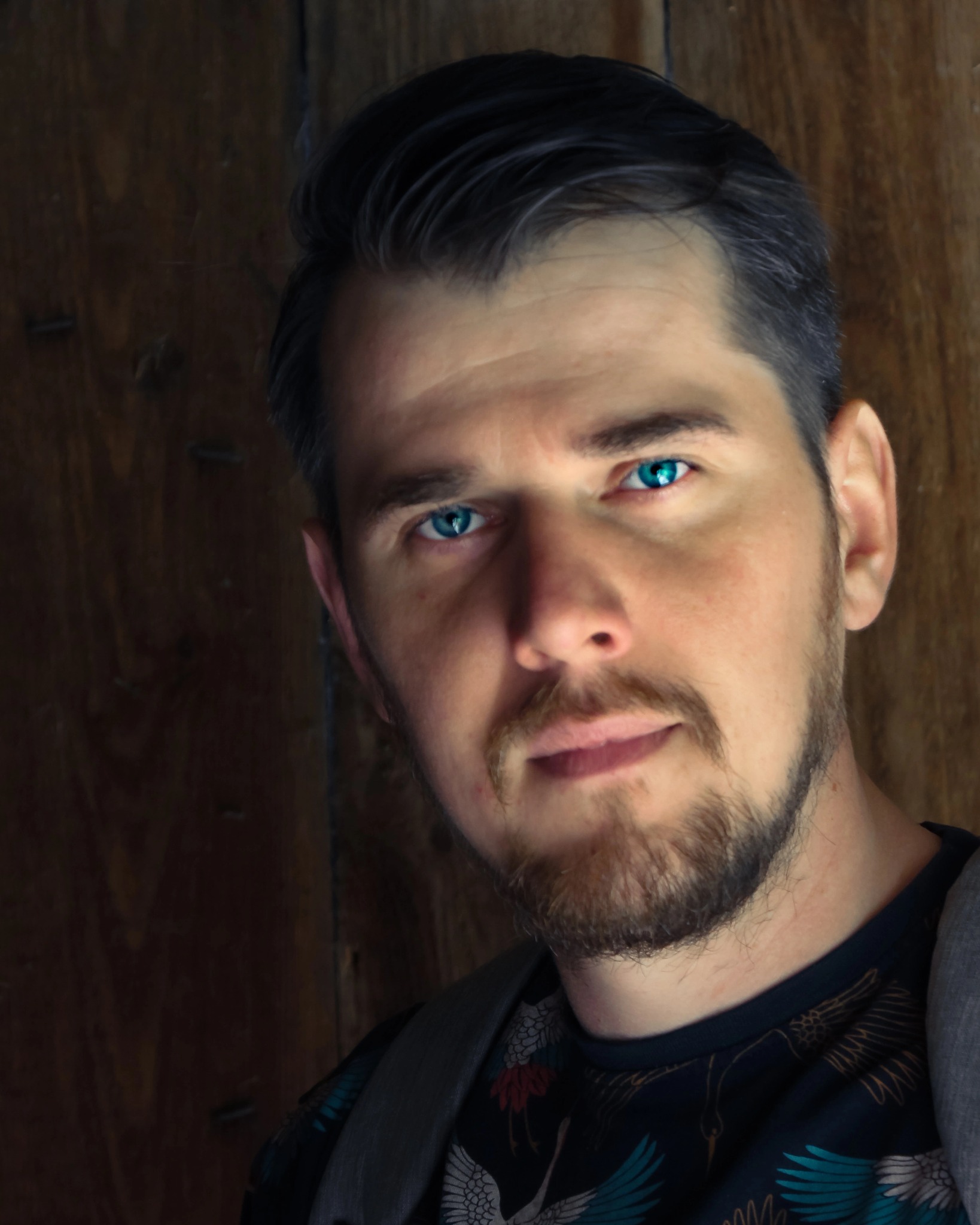 Portret mężczyzny z niebieskimi oczami, z lekkim uśmiechem i brodą, na tle drewnianej ściany.