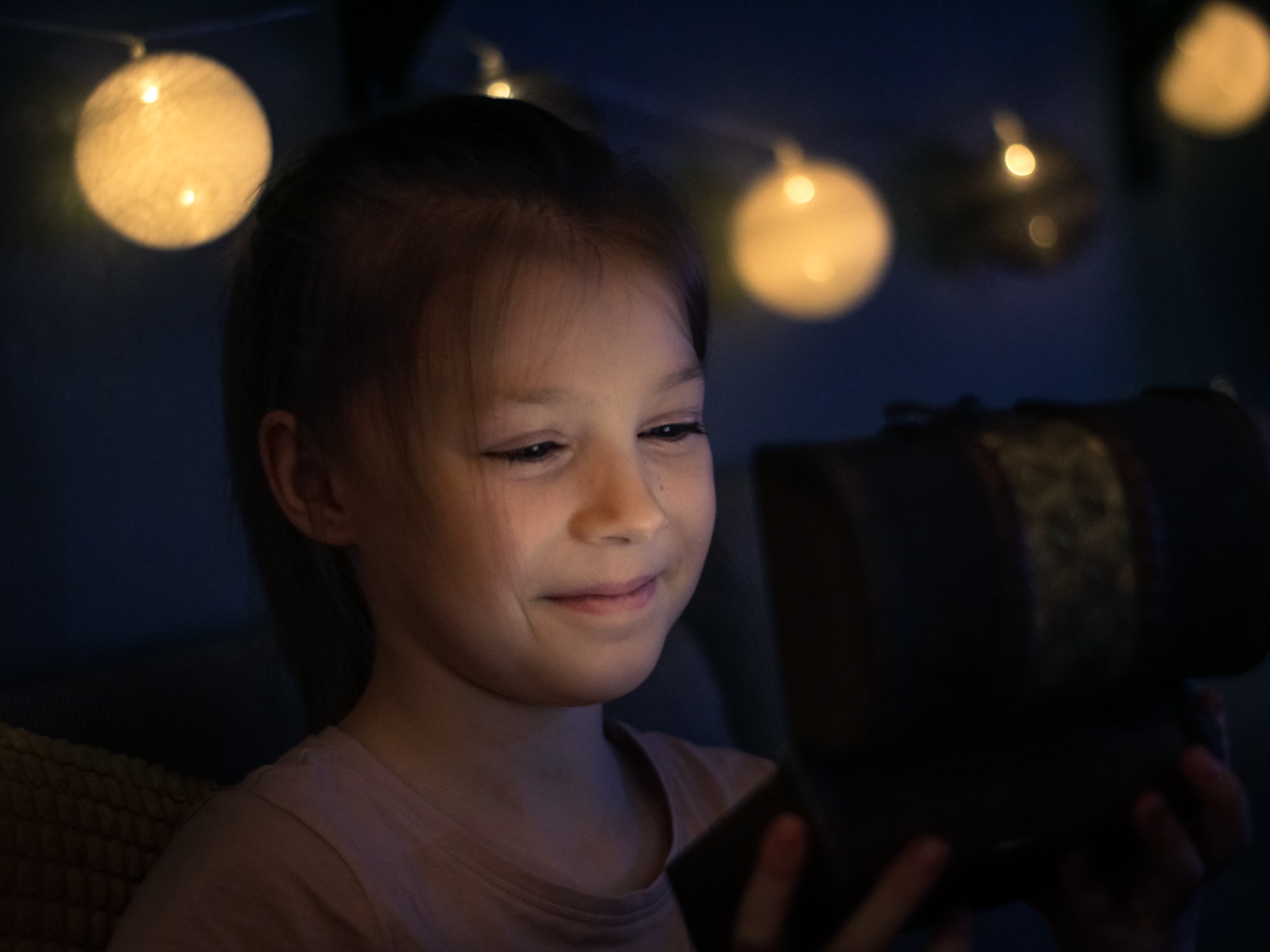 Dziewczynka z zaciekawieniem zagląda do otwartej szkatułki, oświetlona ciepłym światłem girlandy żarówkowej w tle.