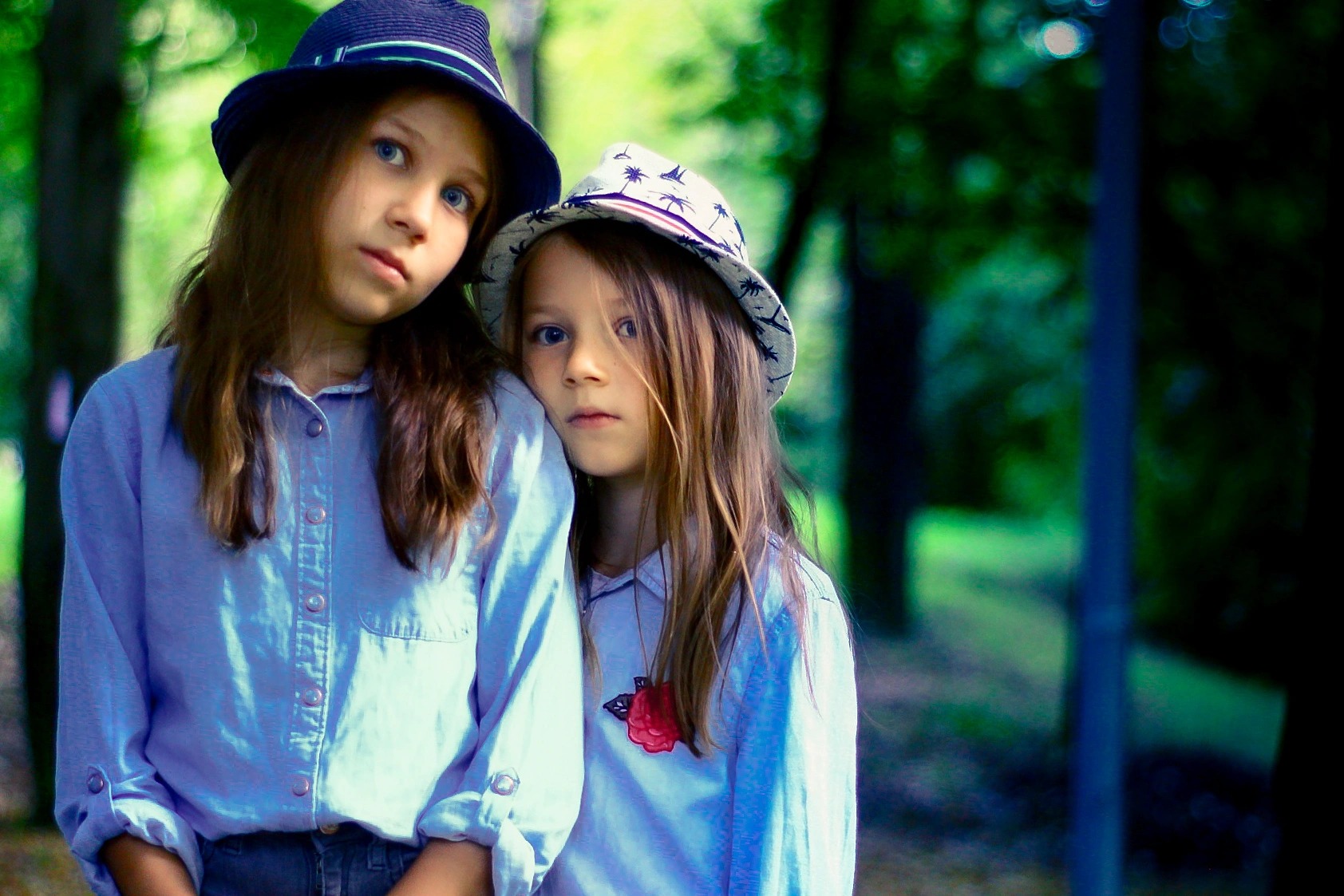 Dwie dziewczynki w kapeluszach, opierające się o siebie w plenerze, z zielonym tłem drzew.