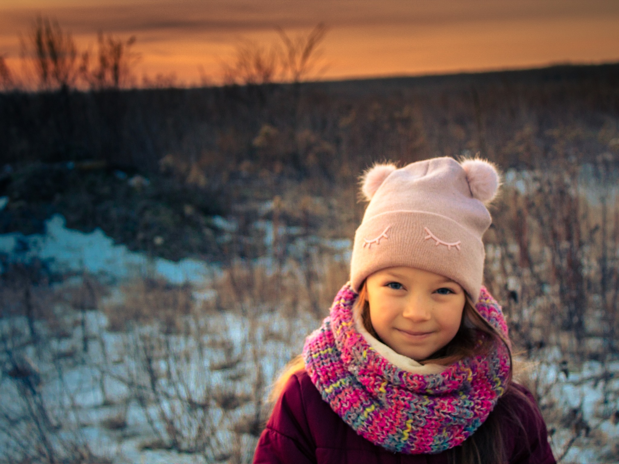 Portret uśmiechniętej dziewczynki w różowej czapce z pomponami i kolorowym szaliku na tle zimowego krajobrazu o zmierzchu.