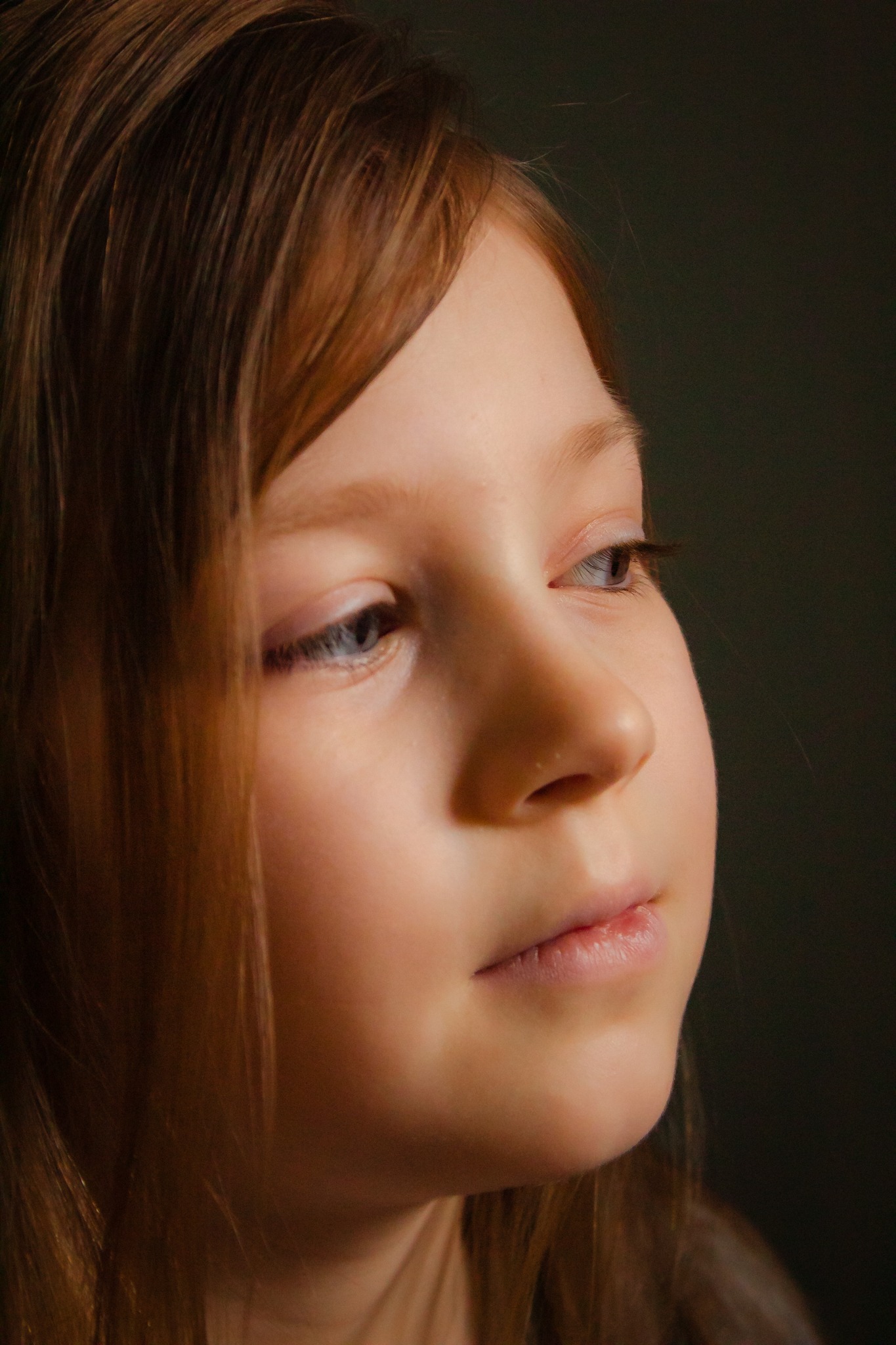 Portret z profilu dziewczynki o jasnej karnacji i długich, rudawych włosach, z delikatnym uśmiechem i błękitnymi oczami, spoglądającej w lewą stronę, na ciemnym, rozmytym tle.