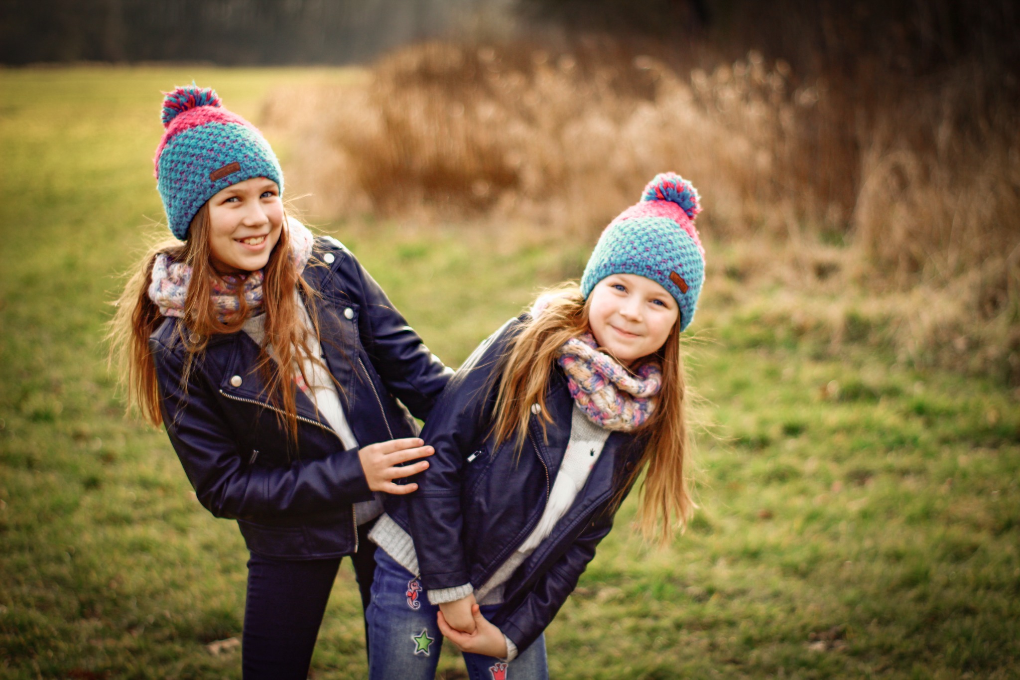 Dwie uśmiechnięte dziewczynki w identycznych, kolorowych czapkach i skórzanych kurtkach pozują na tle łąki z suchą trawą.