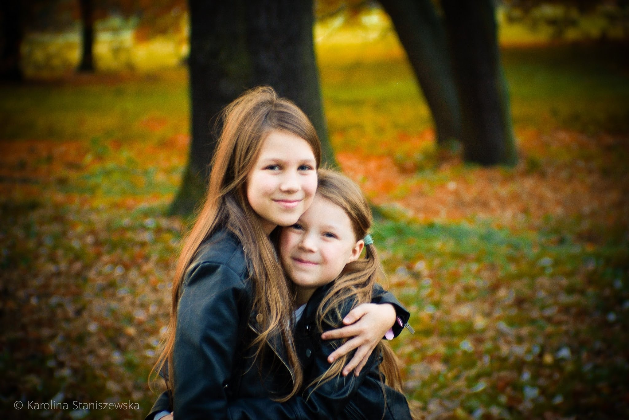 Portret dwóch uśmiechniętych dziewczynek w czarnych kurtkach, obejmujących się na tle jesiennego parku z opadłymi liśćmi.