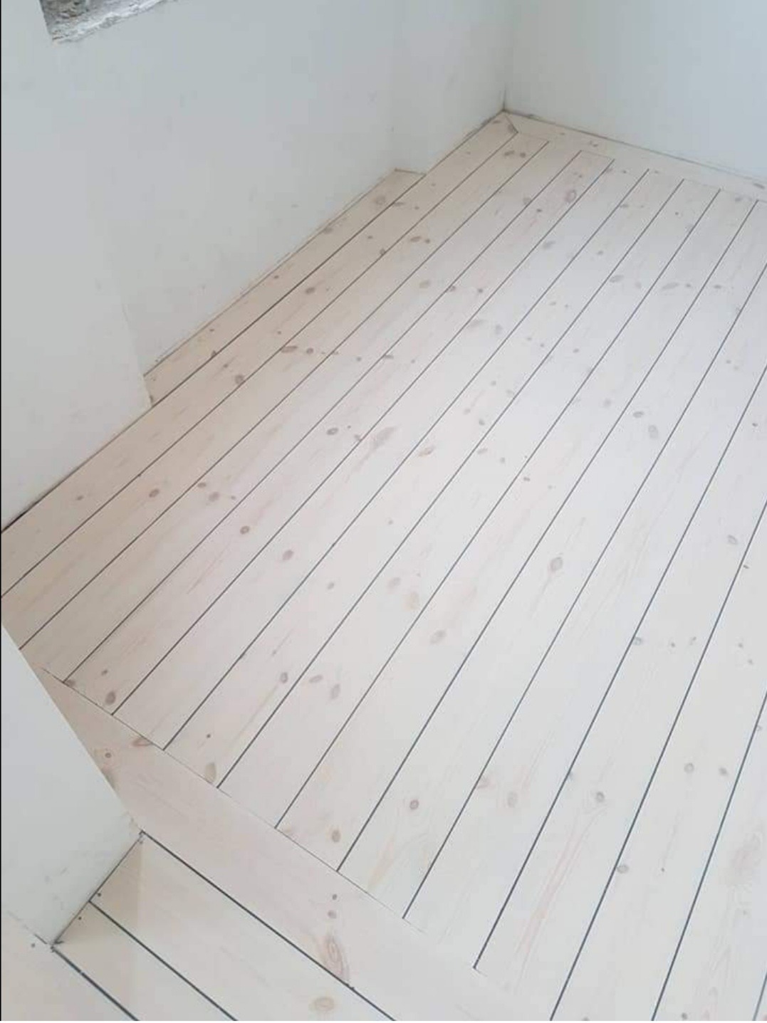 Jasne, bielone deski podłogowe ułożone w nowym pomieszczeniu, widoczne ślady po sękach i delikatne usłojenie drewna, ściany pomalowane na biało.