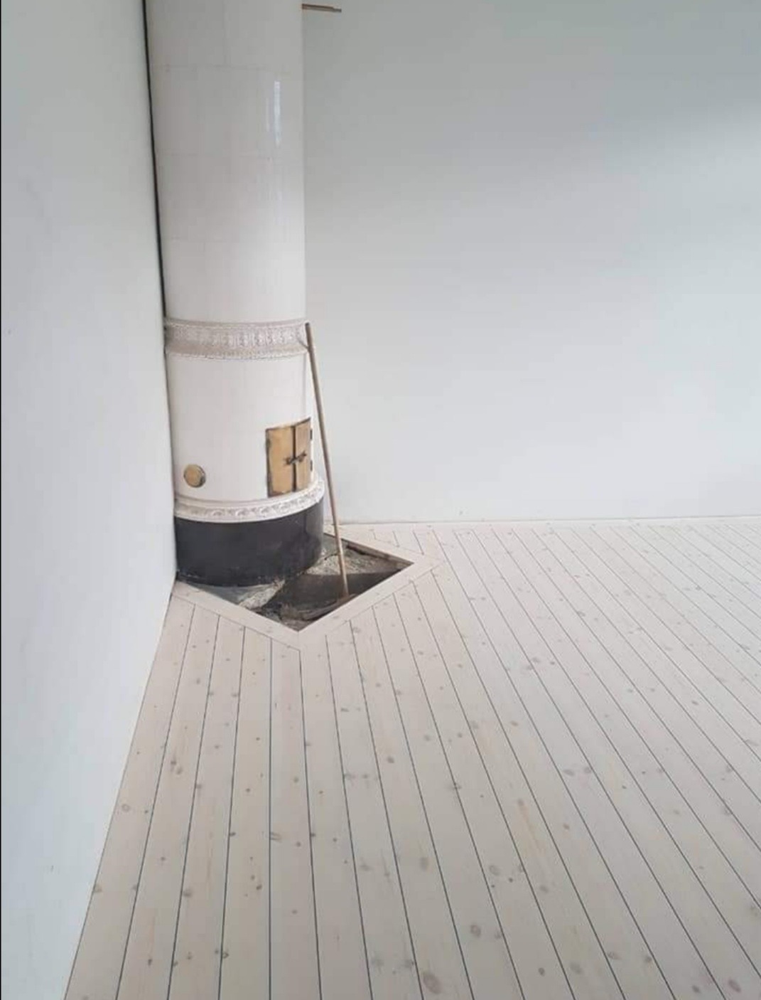 Jasne, drewniane panele podłogowe ułożone wokół podstawy pieca kaflowego z widocznym otworem w podłodze, minimalistyczne wnętrze z białymi ścianami.