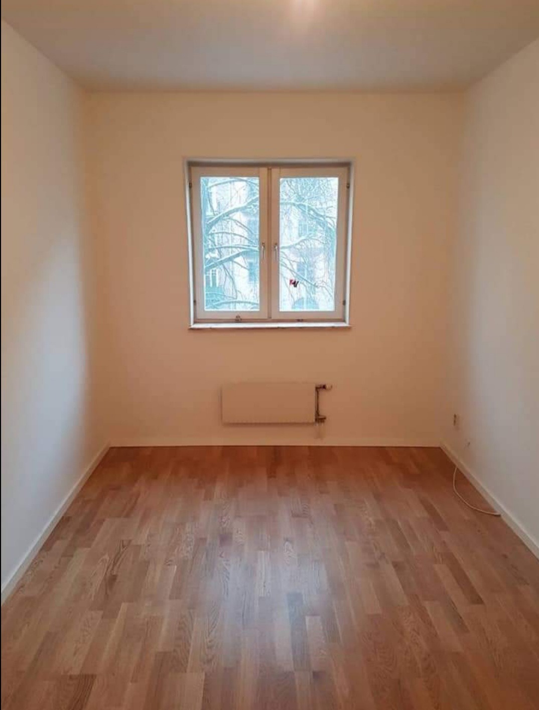 Pusty pokój po remoncie z drewnianą podłogą z paneli, kremowymi ścianami, białym grzejnikiem i oknem z widokiem na drzewa bez liści.