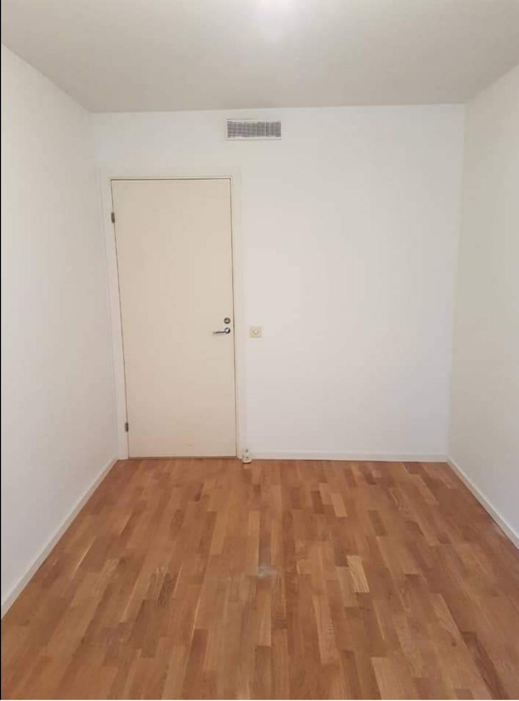 Pusta, jasna przestrzeń z parkietem ułożonym w jodełkę, białe ściany i drzwi, kratka wentylacyjna pod sufitem, gniazdko elektryczne przy podłodze.