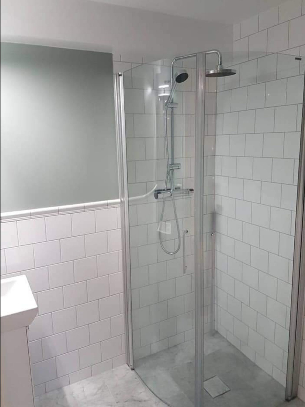 Nowoczesna kabina prysznicowa z deszczownicą i baterią termostatyczną, wykończona białymi, prostokątnymi płytkami, z szarą ścianą w tle i marmurową posadzką.