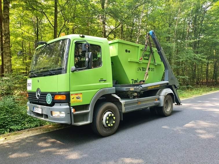 Zielona ciężarówka z hakiem i kontenerem na gruz, zaparkowana na asfalcie na tle lasu.