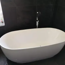 Wolnostojąca wanna owalna, biała, z chromowaną baterią, na tle ciemnych, grafitowych płytek ściennych i podłogowych. Na parapecie okiennym widoczne kosmetyki i środki czystości.