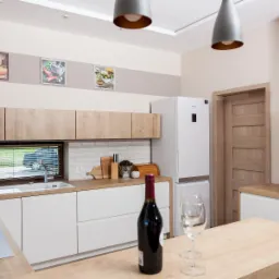 Jasna kuchnia z drewnianymi akcentami, białymi szafkami i sprzętami, ozdobiona butelką wina i kieliszkami na blacie