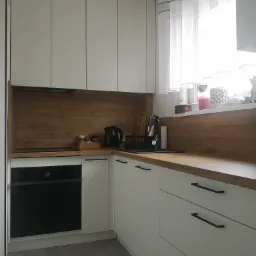 Nowoczesna kuchnia z białymi szafkami, drewnianym blatem i szarymi płytkami podłogowymi. Widoczny czarny piekarnik, czajnik elektryczny i okno z firankami.