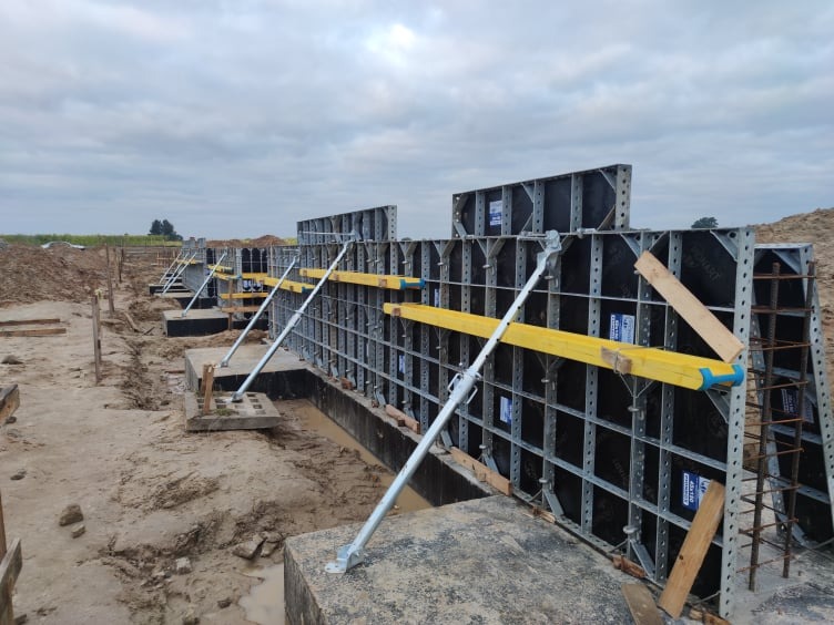 Szczegółowe ujęcie szalunków fundamentowych z widocznym zbrojeniem, wspartych metalowymi stemplami i poziomnicami, na placu budowy w pochmurny dzień.