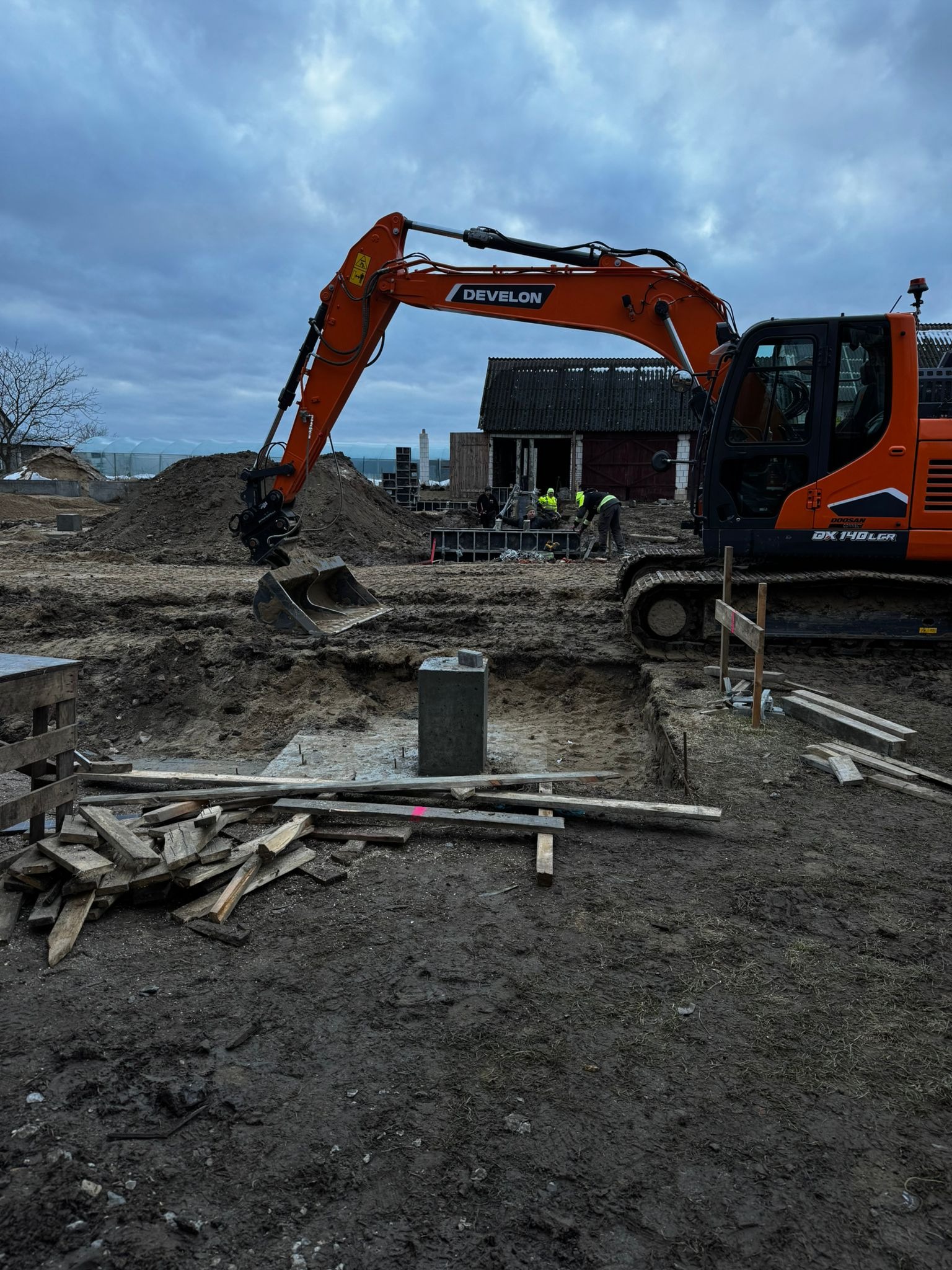 Pomarańczowa koparka Develon na placu budowy, wykop pod fundamenty, widoczne szalunki i zbrojenie. W tle budynek gospodarczy i pracownicy w żółtych kamizelkach.