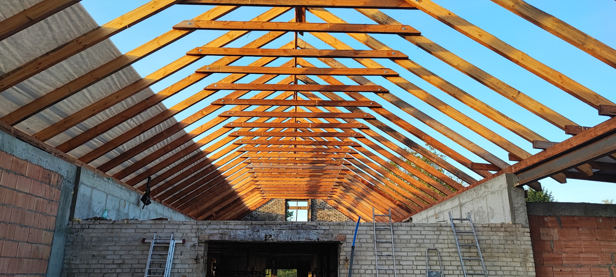 Konstrukcja drewniana dachu w trakcie budowy, widok od wewnątrz. Elementy więźby dachowej na tle błękitnego nieba. Ściany z cegły i betonu, drabiny budowlane.
