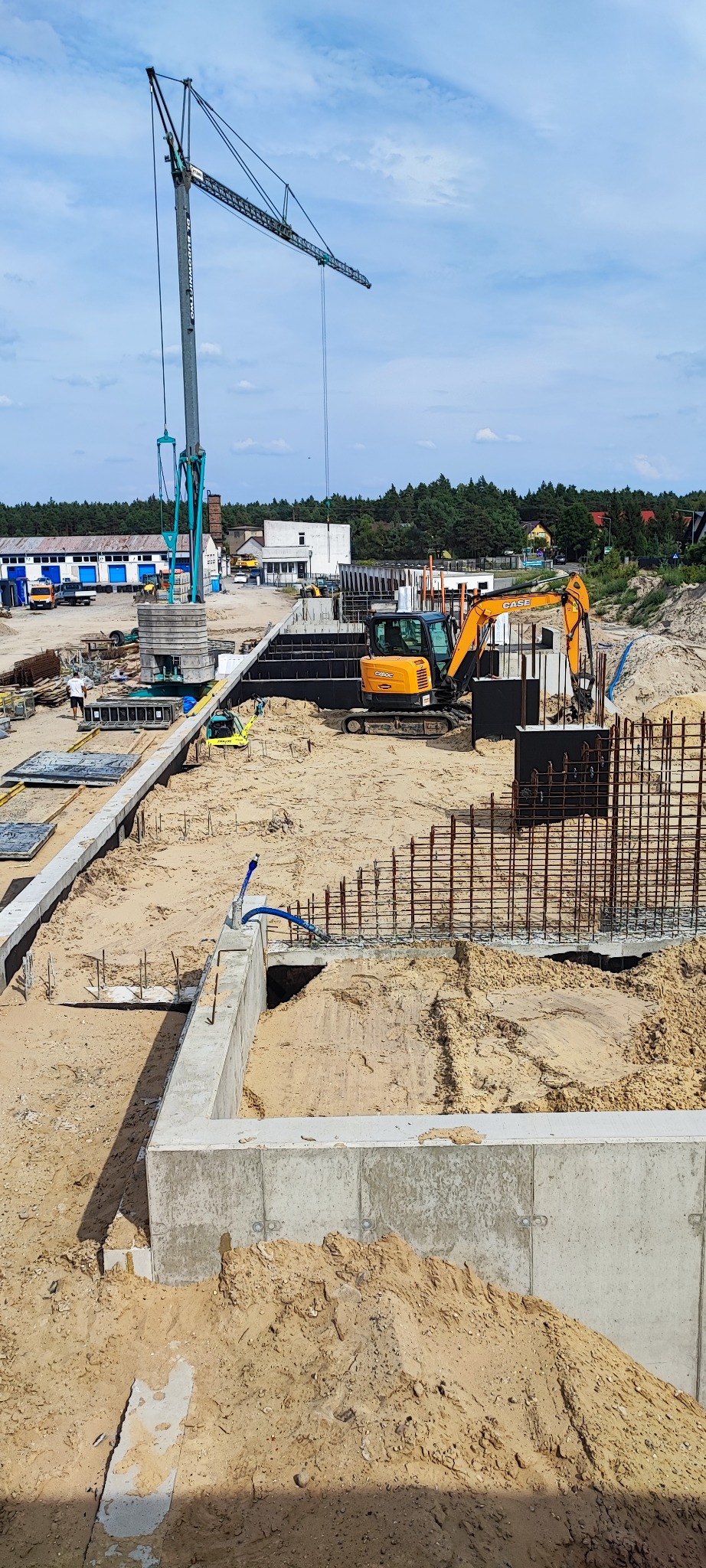 Fundamenty domu w trakcie budowy z widocznym zbrojeniem, szalunkiem i koparką. Na drugim planie dźwig budowlany i budynki gospodarcze.