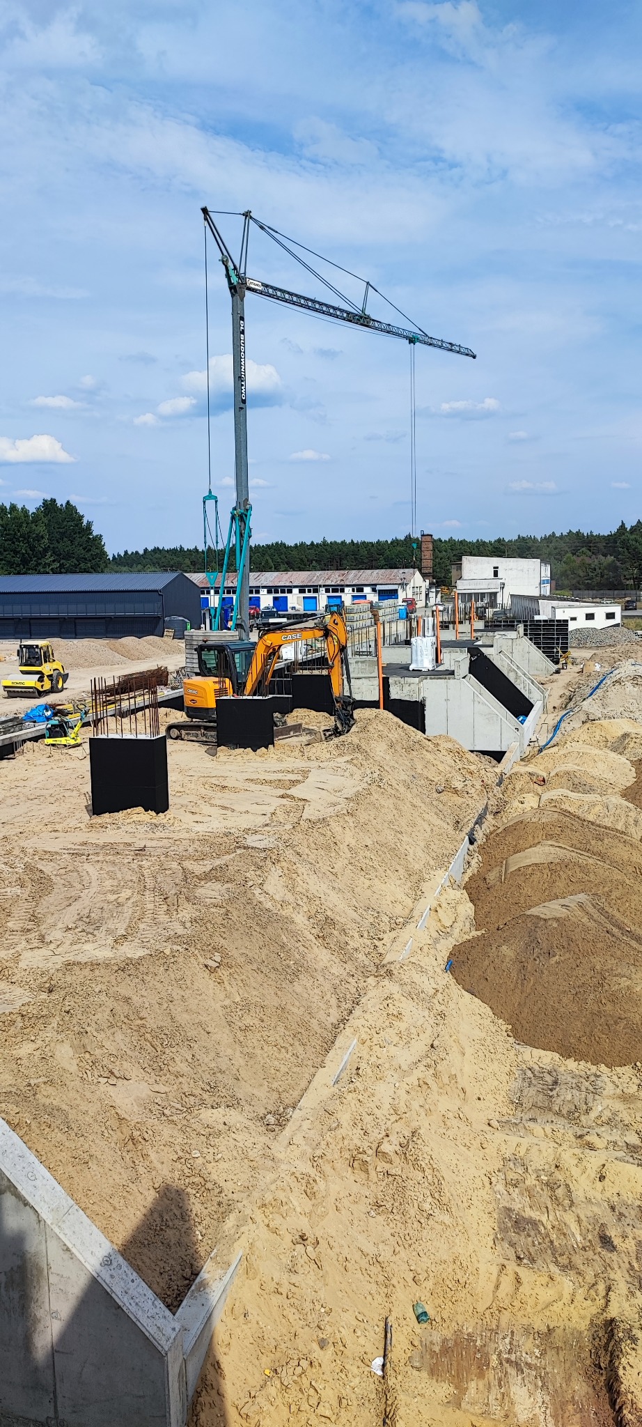 Budowa fundamentów z użyciem dźwigu i koparki na placu budowy. Widoczne zbrojenia, betonowe elementy i przygotowany teren pod dalsze prace. Słoneczny dzień.