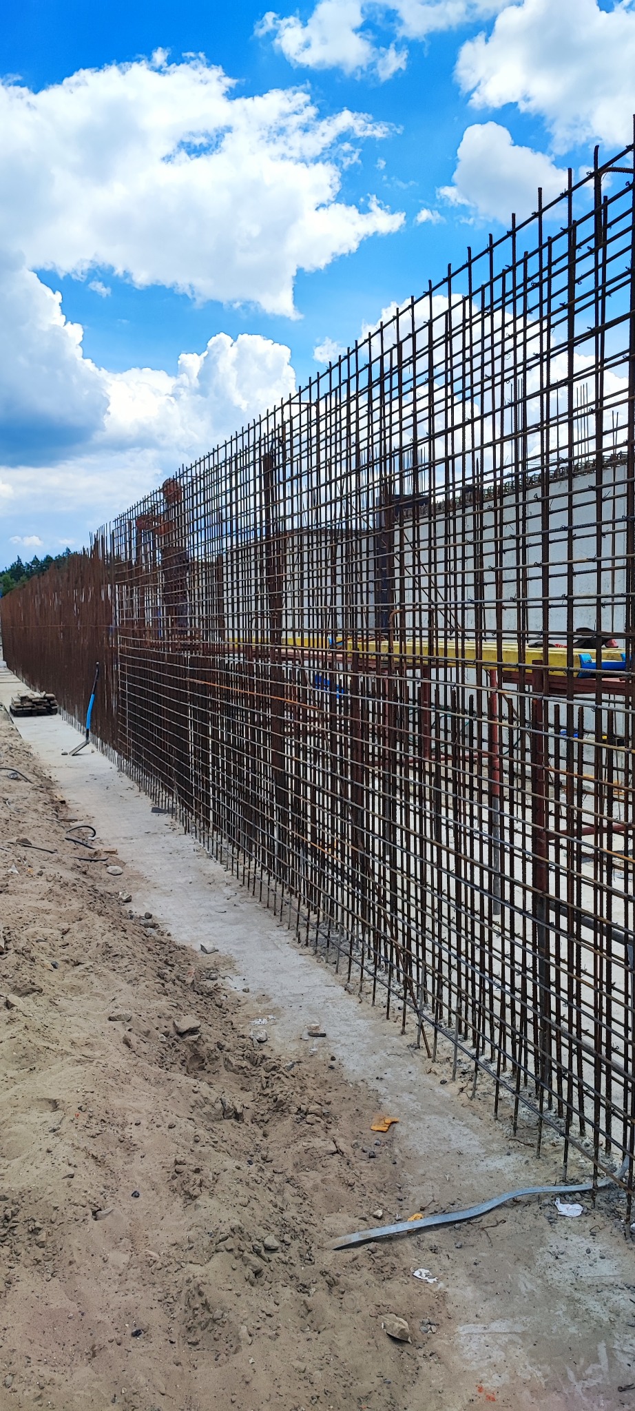 Zbrojenie fundamentów pod budowę, widok na siatkę z prętów stalowych na tle błękitnego nieba z chmurami. Detal: widoczne elementy szalunku i podłoże z piasku.