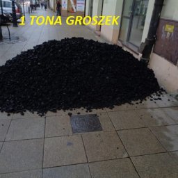 Transopex - Góra czarnego paliwa stałego, prawdopodobnie ekogroszku, wysypana na chodniku przed budynkiem z rynną spustową i włazem kanalizacyjnym w pobliżu, z napisem '1 TONA groszek' nałożonym na zdjęcie.