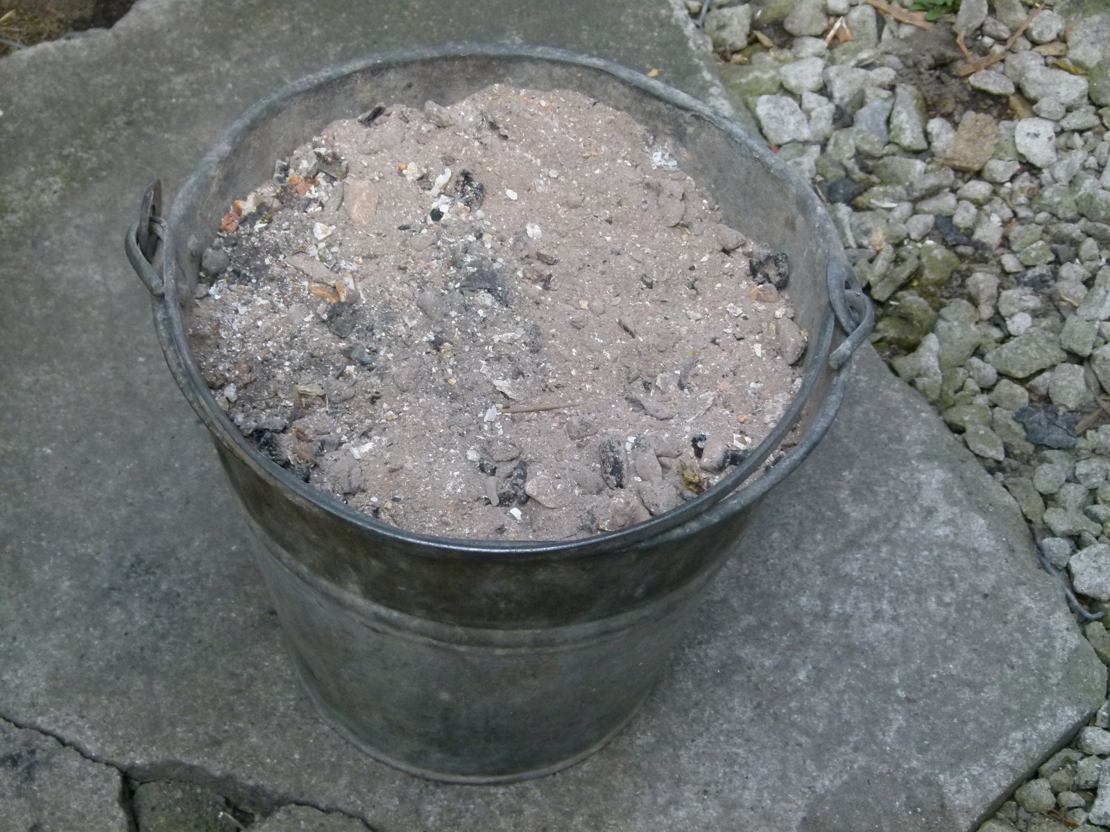 Metalowe wiadro wypełnione popiołem i niedopałkami, stojące na betonowej płycie, obok której leżą drobne kamienie.