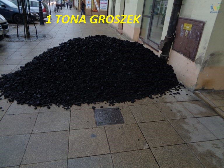 Góra czarnego paliwa stałego, prawdopodobnie ekogroszku, wysypana na chodniku przed budynkiem z rynną spustową i włazem kanalizacyjnym w pobliżu, z napisem '1 TONA groszek' nałożonym na zdjęcie.